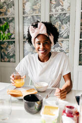 Positive schwarze Frau mit einem Glas frischem Saft, die fröhlich lächelt und wegschaut, während sie beim Frühstück zu Hause am Tisch sitzt - ADSF39421