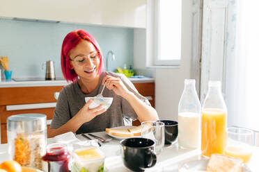 Optimistic young woman with dyed hair smiling and enjoying healthy dish while sitting at table in cozy kitchen at home in morning - ADSF39423
