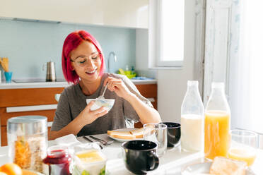 Optimistische junge Frau mit gefärbten Haaren, die lächelt und ein gesundes Gericht genießt, während sie am Tisch in einer gemütlichen Küche zu Hause am Morgen sitzt - ADSF39423