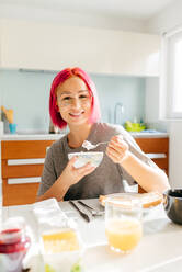 Optimistische junge Frau mit gefärbten Haaren, die lächelt und ein gesundes Gericht genießt, während sie am Tisch in einer gemütlichen Küche zu Hause am Morgen sitzt - ADSF39424