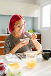 Optimistische junge Frau mit gefärbten Haaren, die lächelt und ein gesundes Gericht genießt, während sie am Tisch in einer gemütlichen Küche zu Hause am Morgen sitzt - ADSF39425