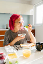 Optimistische junge Frau mit gefärbten Haaren, die lächelt und ein gesundes Gericht genießt, während sie am Tisch in einer gemütlichen Küche zu Hause am Morgen sitzt - ADSF39426