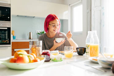 Optimistische junge Frau mit gefärbten Haaren, die lächelt und ein gesundes Gericht genießt, während sie am Tisch in einer gemütlichen Küche zu Hause am Morgen sitzt - ADSF39427