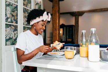 Happy African American lady smiling and adding butter on toast while sitting at table and having breakfast at home - ADSF39430