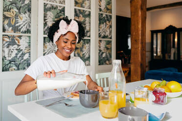 African american woman smiling and pouring milk on bowl while having breakfast in morning at home - ADSF39432