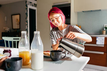 Young woman with dyed hair smiling and filling mug with fresh coffee while having breakfast in morning at home - ADSF39433