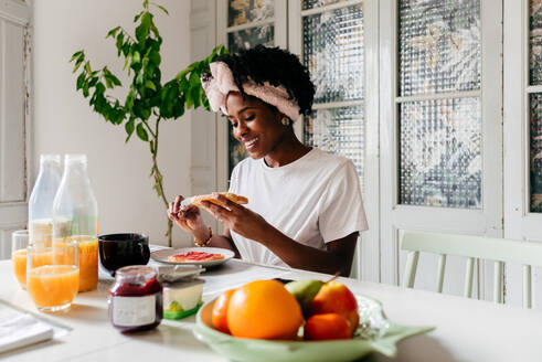 Afroamerikaner, der Marmelade auf frischen Toast streicht, während er am Tisch sitzt und zu Hause frühstückt - ADSF39434