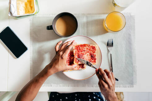 Draufsicht auf eine anonyme afroamerikanische Person, die Marmelade auf frischen Toast streicht, während sie am Tisch sitzt und zu Hause frühstückt - ADSF39435