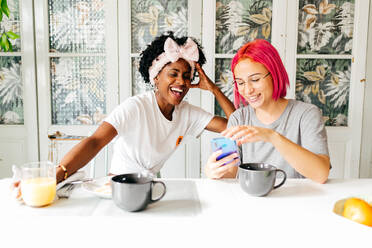 Junge Frau mit rosafarbenen Haaren, die ihrem fröhlichen afroamerikanischen Freund beim gemeinsamen Frühstück zu Hause lächelnd ihr Smartphone zeigt - ADSF39438