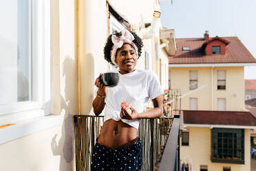 Entspannte afroamerikanische Frau mit Toast, die auf dem Balkon steht und ein heißes Getränk am Morgen schlürft - ADSF39444