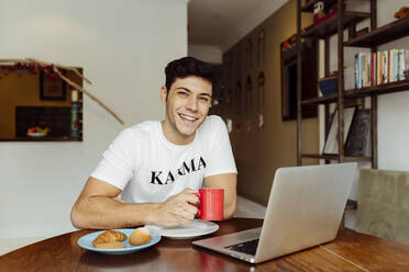 Handsome caucasian man having breakfast at home while working on his laptop on the table - ADSF39446