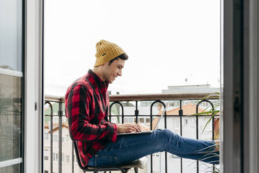 Seitenansicht eines trendigen, konzentrierten kaukasischen Mannes mit Mütze und Hemd, der auf einem Stuhl auf dem Balkon sitzt, während er zu Hause an seinem Laptop arbeitet - ADSF39448