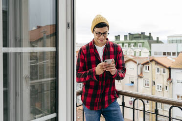 Attractive hipster caucasian man with beanie hat an glasses standing on balcony at home using mobile phone - ADSF39450