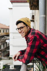 Trendy handsome cheerful caucasian man with beanie hat, shirt and glasses leaning on fence of a balcony at home looking away - ADSF39451