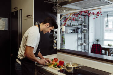 Seitenansicht eines gut aussehenden kaukasischen Mannes mit Schürze, der in der Küche zu Hause Gemüse schneidet - ADSF39453