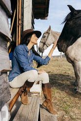 Schöne Brünette in Hut steht in der Tür der Ranch Stall mit Pferden in der Nähe lächelnd im Sonnenlicht - ADSF39457