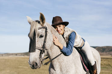 Seitenansicht einer Frau mit Hut, die auf einem schönen weißen Pferd vor dem Hintergrund einer ländlichen Landschaft unter blauem Himmel sitzt und in die Kamera schaut - ADSF39461