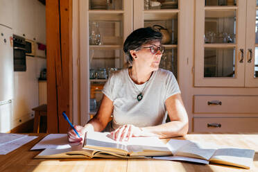 Fokussierte Frau mittleren Alters mit Brille und Stift in der Hand sitzt am Tisch mit einem Haufen Bücher und Papiere und studiert professionelle Materialien zu Hause - ADSF39466