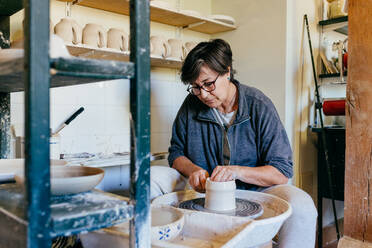 Eine Handwerkerin mittleren Alters formt eine Keramikvase auf der Töpferscheibe, während sie im Atelier arbeitet - ADSF39467