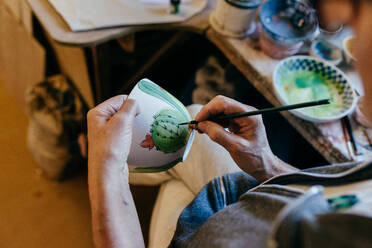 Anonymous focused mature craftswoman with paintbrush painting ceramic bowl while making earthenware in pottery workshop - ADSF39469