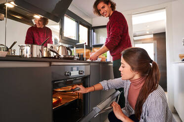 Positive junge Frau in Freizeitkleidung öffnet die Ofentür und kauert neben ihrem lächelnden Freund, der das Geschirr abwäscht, während sie gemeinsam in einer modernen Küche kocht - ADSF39474