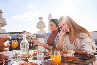 Happy young female friends eating delicious pizza and chatting while gathering on terrace on sunny summer day - ADSF39476