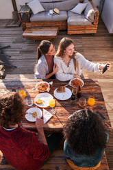 Hohe Winkel junge positive Freunde in Freizeitkleidung genießen leckeres Mittagessen auf der Terrasse und nehmen Selfie auf Smartphone auf sonnigen Sommertag - ADSF39477