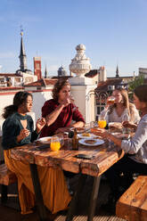 Positiv gestimmte junge Freunde in legeren Outfits genießen ein leckeres Mittagessen am Esstisch auf der sonnigen Terrasse - ADSF39478