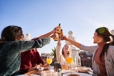 Eine Gruppe zufriedener junger Freunde stößt mit Flaschen kalten Biers an, während sie auf einer sonnigen Terrasse ein leckeres Mittagessen zu sich nehmen - ADSF39479