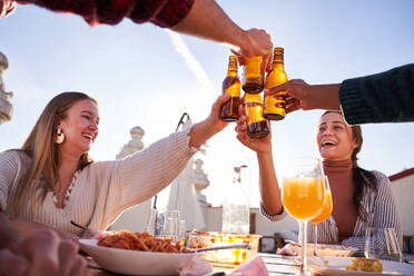 Eine Gruppe zufriedener junger Freunde stößt mit Flaschen kalten Biers an, während sie auf einer sonnigen Terrasse ein leckeres Mittagessen zu sich nehmen - ADSF39480