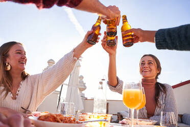 Eine Gruppe zufriedener junger Freunde stößt mit Flaschen kalten Biers an, während sie auf einer sonnigen Terrasse ein leckeres Mittagessen zu sich nehmen - ADSF39481