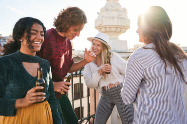 Lachende junge gemischtrassige Freunde in Freizeitkleidung genießen kaltes Bier und tanzen fröhlich bei einer Party auf dem Dach bei sonnigem Wetter - ADSF39483