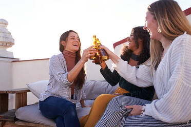 Unbeschwerte junge multiethnische Freundinnen in legerer Kleidung stoßen mit Flaschen kalten Biers an, während sie auf einem gemütlichen Sofa auf einer sonnigen Terrasse sitzen - ADSF39484