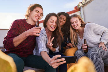 Gruppe aufgeregter junger multirassischer Freunde in Freizeitkleidung, die Grimassen schneiden und ein Selfie mit ihrem Smartphone machen, während sie sich mit Bierflaschen auf einer sonnigen Terrasse versammeln - ADSF39486