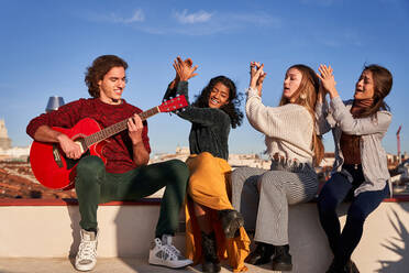 Fröhliche multiethnische Frauen, die in die Hände klatschen und singen, während sie neben einem lächelnden jungen Mann sitzen, der auf einer sonnigen Terrasse Akustikgitarre spielt - ADSF39487