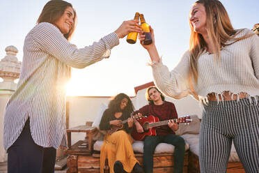 Glückliche junge Frauen stoßen mit kalten Bierflaschen an, während sie auf einem sonnigen Dach in der Nähe verschiedener Freunde stehen, die auf dem Sofa Gitarre und Ukulele spielen - ADSF39488