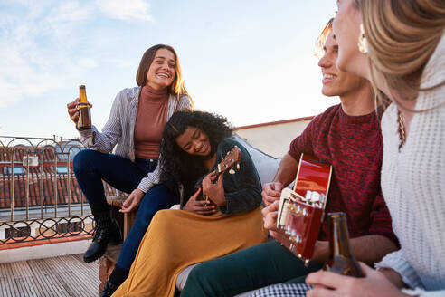Fröhliche junge gemischtrassige Freunde in Freizeitkleidung spielen Akustikgitarre und Ukulele, während sie sich gemeinsam auf einem bequemen Sofa auf dem Dach eines Gebäudes ausruhen - ADSF39489