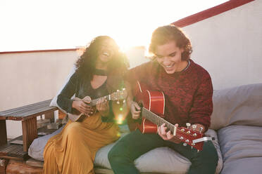 Excited young multiethnic friends in casual wear playing acoustic guitar and ukulele while sitting together on comfy couch on sunny rooftop - ADSF39490