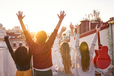 Rückansicht gesichtslose aufgeregte Freunde mit erhobenen Armen im Rücken, die Freizeitkleidung tragen und den Sonnenuntergang bewundern, während sie auf einem Dach stehen - ADSF39493