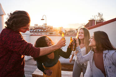 Eine Gruppe zufriedener junger Freunde stößt auf einer sonnigen Terrasse auf Flaschen mit kaltem Bier an - ADSF39495