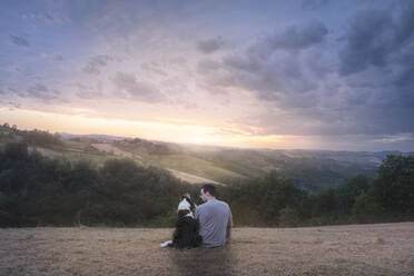 Ein Mann und sein Border Collie Hund sitzen auf einem Feld in der Landschaft und genießen den Sonnenuntergang, Italien, Europa - RHPLF23292