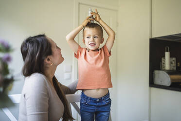 Niedlicher Junge balanciert zu Hause bei seiner Mutter eine Farbtube auf dem Kopf - DMMF00170