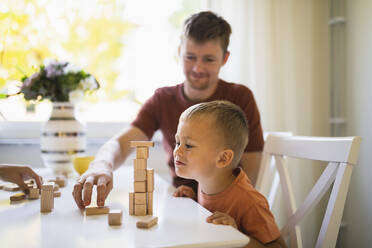 Cute boy looking at father stacking toy blocks on dining table - DMMF00177