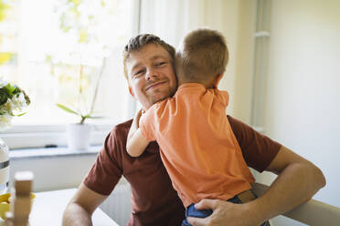Junge umarmt lächelnden Vater zu Hause - DMMF00179