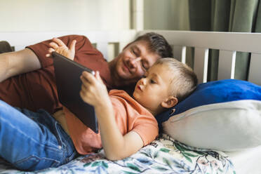 Sohn und Vater sehen sich zu Hause auf dem Bett liegend einen Tablet-PC an - DMMF00186