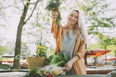 Smiling woman showing bunch of carrots at local market - NDEF00007