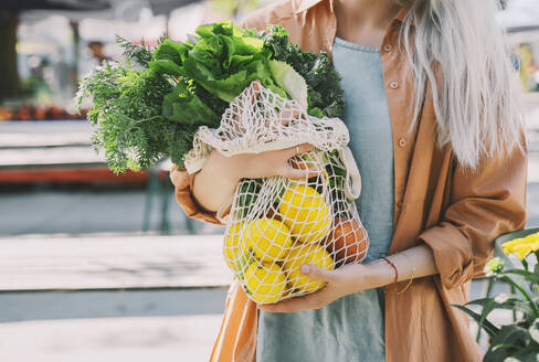 Customer holding leafy vegetables and lemons in mesh bag at local market - NDEF00009