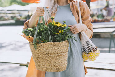 Woman holding flowers and vegetables in wicker bag at local market - NDEF00011