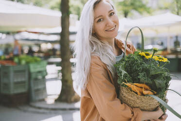 Lächelnde Frau mit Blumen und Gemüse in einer wiederverwendbaren Korbtasche auf dem Markt - NDEF00012