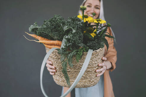 Woman showing wicker bag filled with vegetables and flowers against gray background - NDEF00017